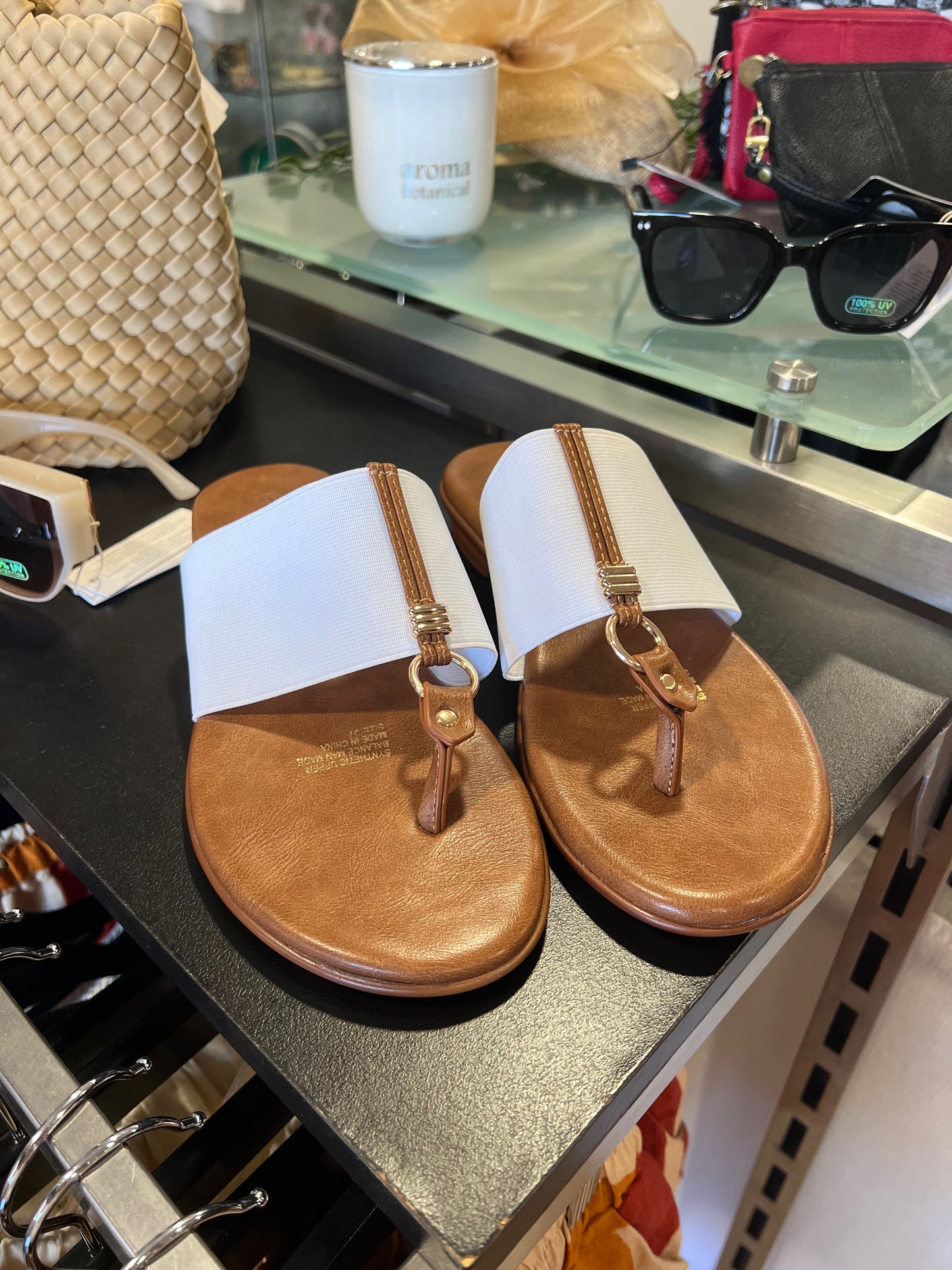A pair of white sandals with brown straps and a label attached, displayed on a glass shelf in a store.