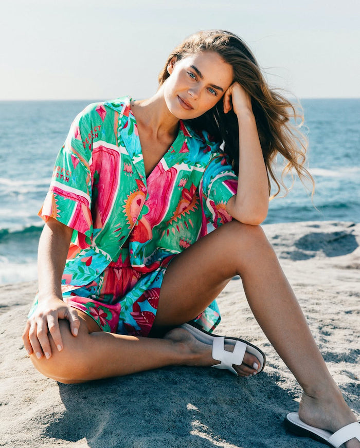 A woman sitting on a rocky surface by the beach, wearing a short set with a floral print, consisting of a shirt and shorts, in a casual, relaxed pose.