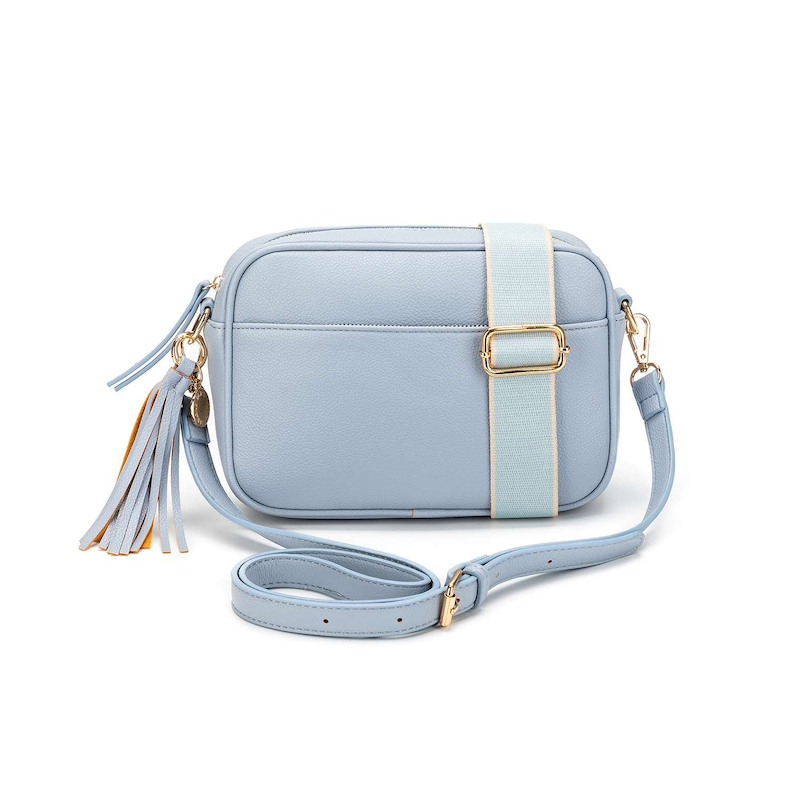 A blue chambray crossbody bag with three adjustable straps, a tassel detail, and a brushed gold hardware closure.