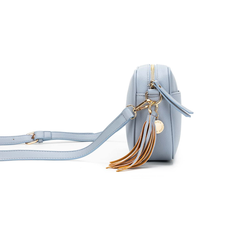 A blue chambray crossbody bag with three adjustable straps, a tassel detail, and a brushed gold hardware closure.