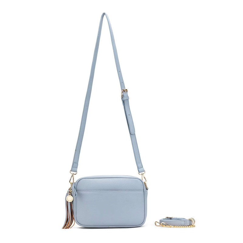 A blue chambray crossbody bag with three adjustable straps, a tassel detail, and a brushed gold hardware closure.