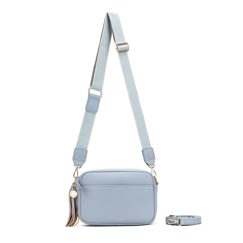 A blue chambray crossbody bag with three adjustable straps, a tassel detail, and a brushed gold hardware closure.