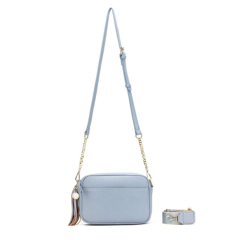A blue chambray crossbody bag with three adjustable straps, a tassel detail, and a brushed gold hardware closure.