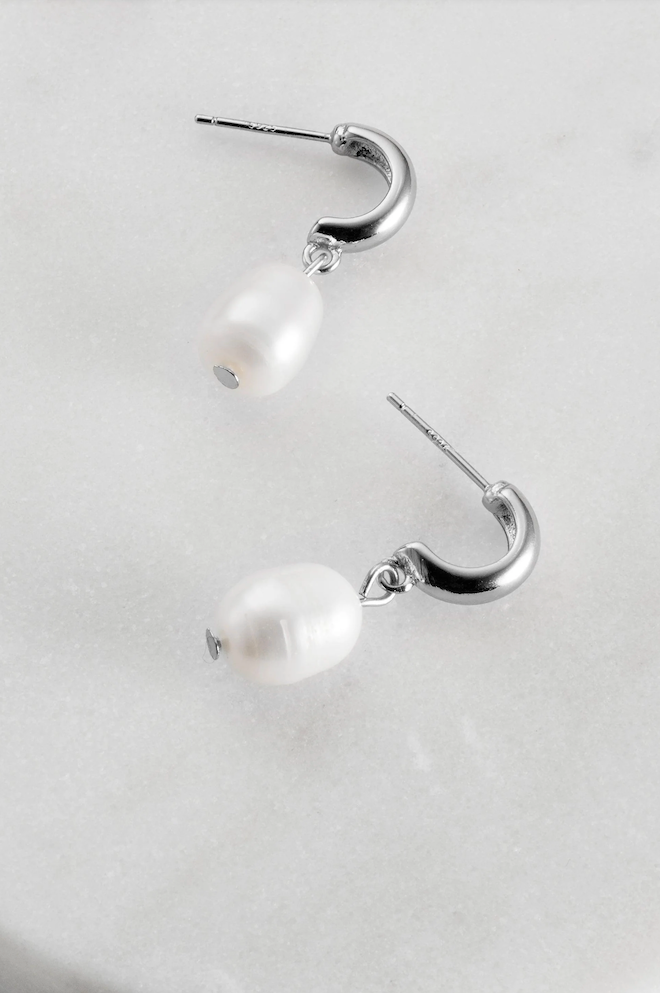 A pair of silver-colored hoop earrings with fresh water pearl adornments on a marble surface.