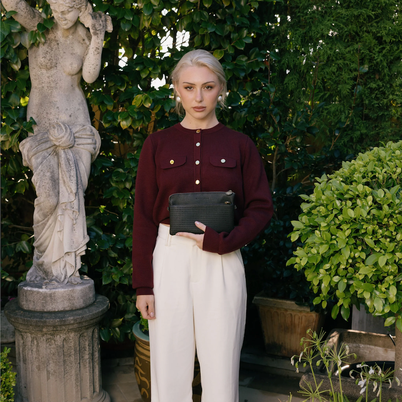 A woman wearing a red cardigan and white trousers is holding a black crossbody clutch with gold hardware.