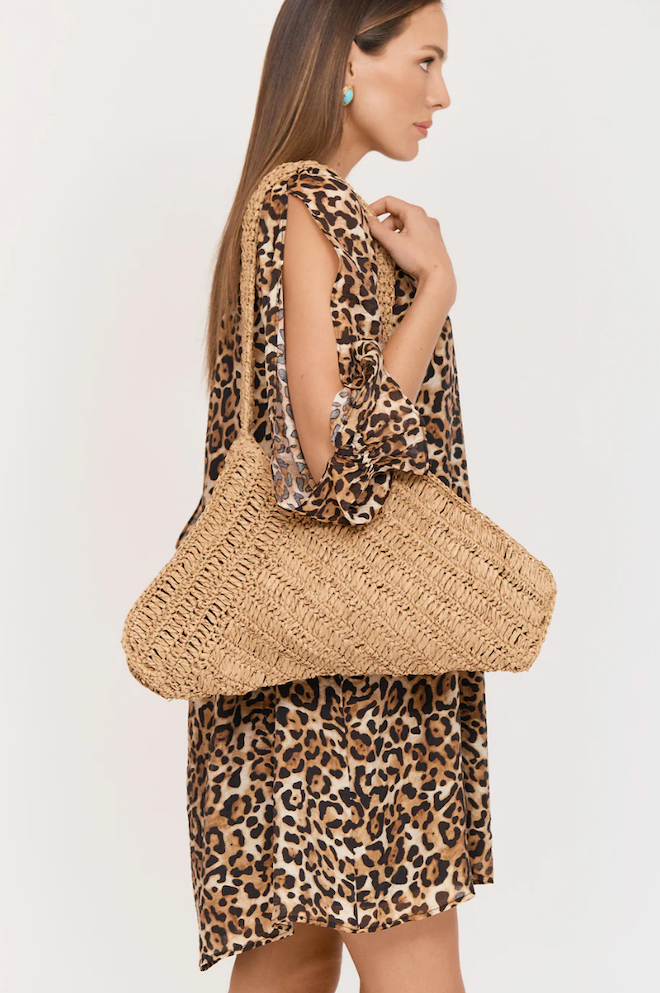 Woman wearing a leopard print dress holding a straw bag on a plain background