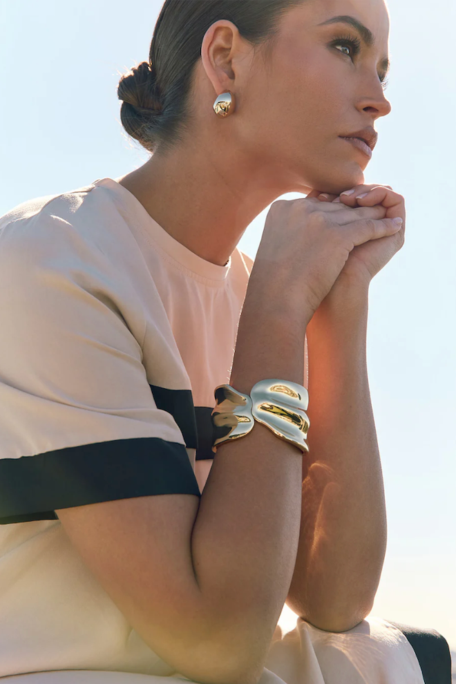 Woman wearing a white top with black trim, gold bracelet, and earrings against a bright background