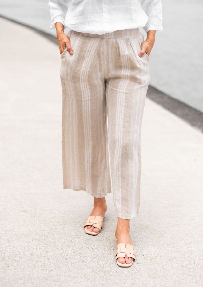 A person standing by a body of water, wearing Valentina Striped Linen Pants with a beige and white stripe pattern, paired with a white sleeveless top.