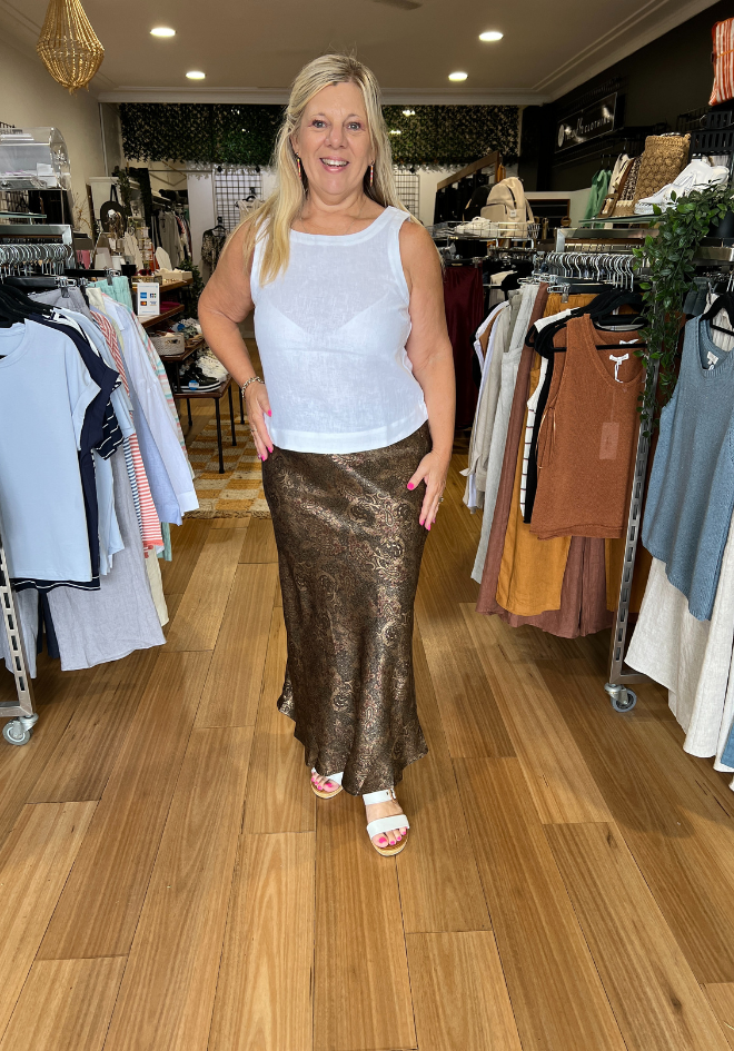 A person standing in a retail environment wearing a white sleeveless top and olive green satin skirt with a paisley pattern and gold accents.