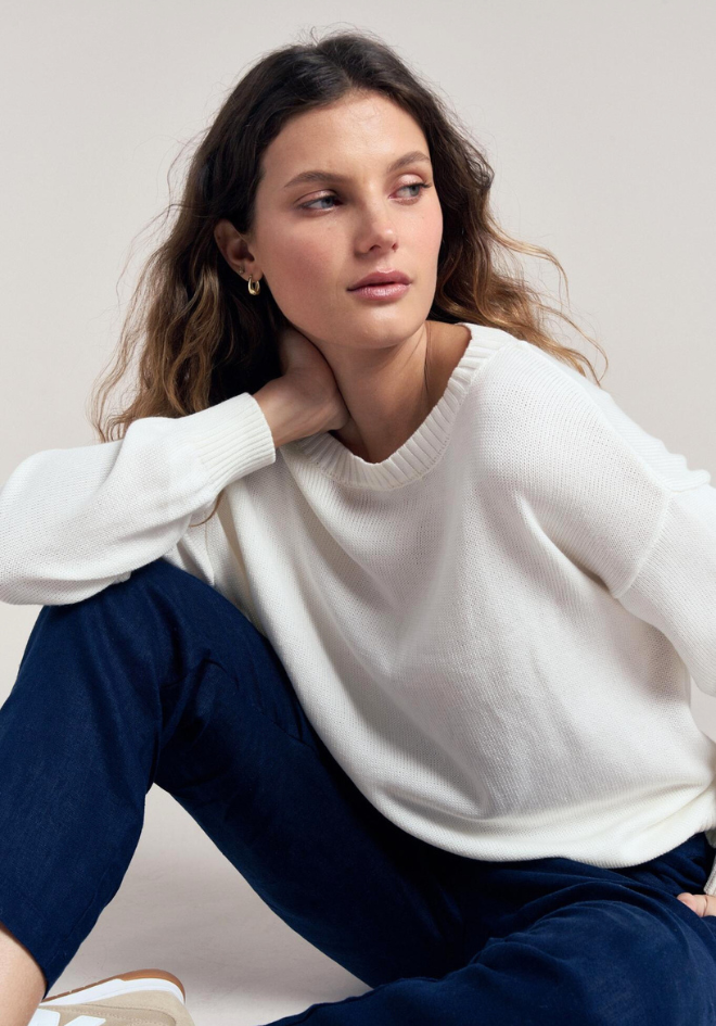 Woman wearing a white sweater and navy pants sitting on a light gray background