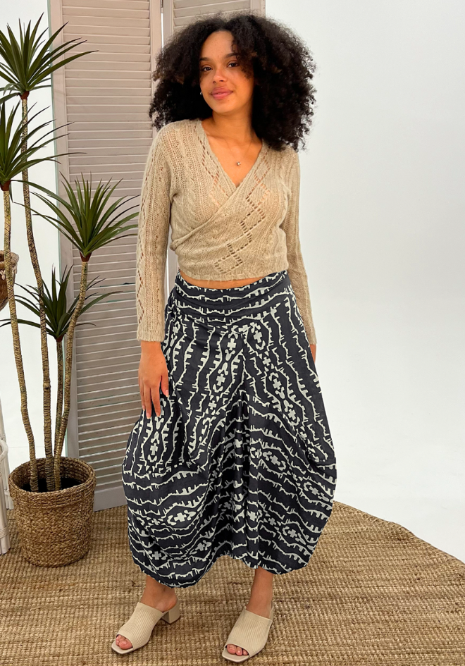 Woman wearing a beige sweater and patterned pants standing indoors with plants in the background