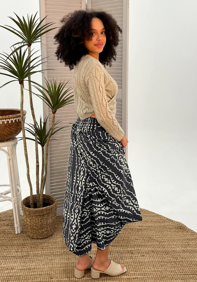 Woman wearing a beige sweater and patterned skirt standing indoors with plants in the background