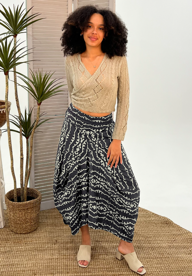 Woman wearing a beige sweater and patterned skirt standing indoors with plants in the background