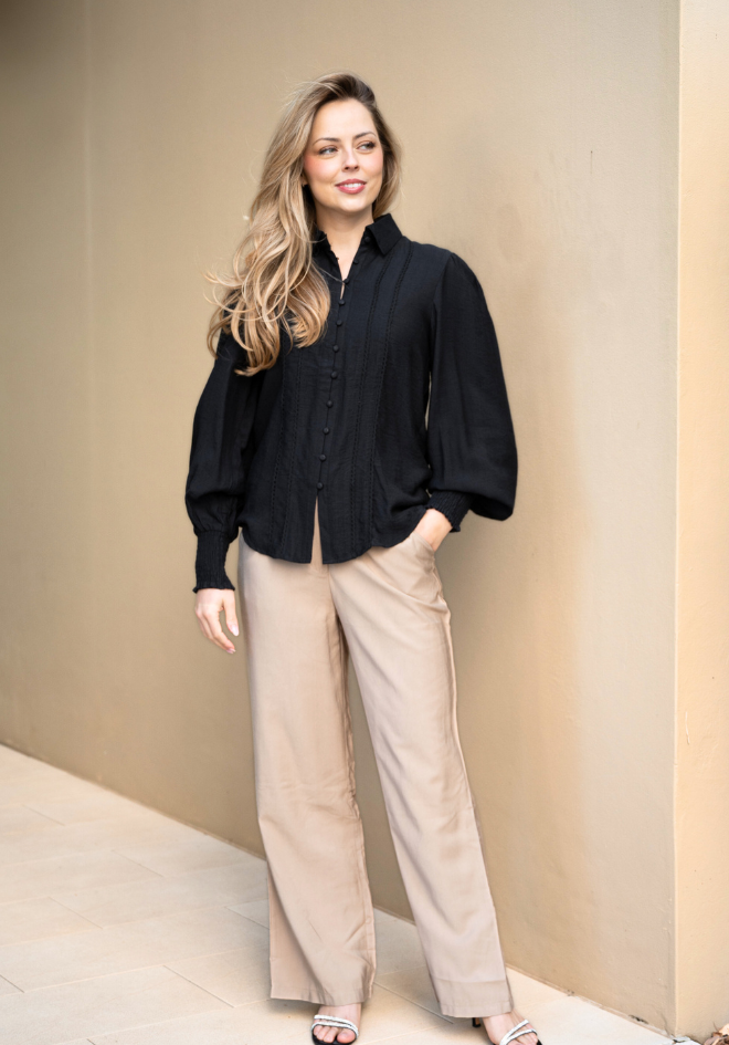 Woman wearing a black blouse and beige pants against a beige wall.