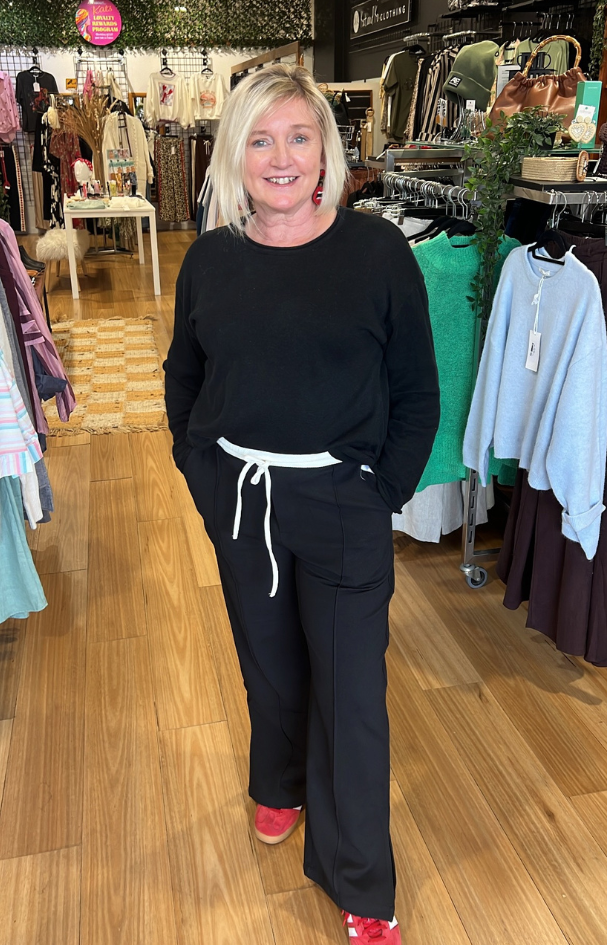 Woman in a black outfit standing in a clothing store with racks of clothes in the background.