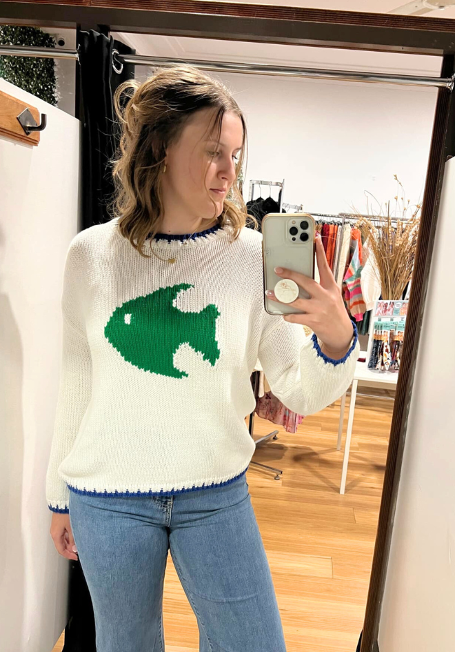 Person wearing a white sweater with a green fish design, taking a mirror selfie.