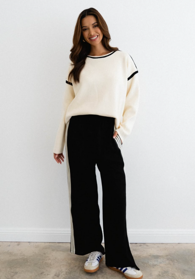 Woman wearing a cream sweater with black trim and black pants against a white background