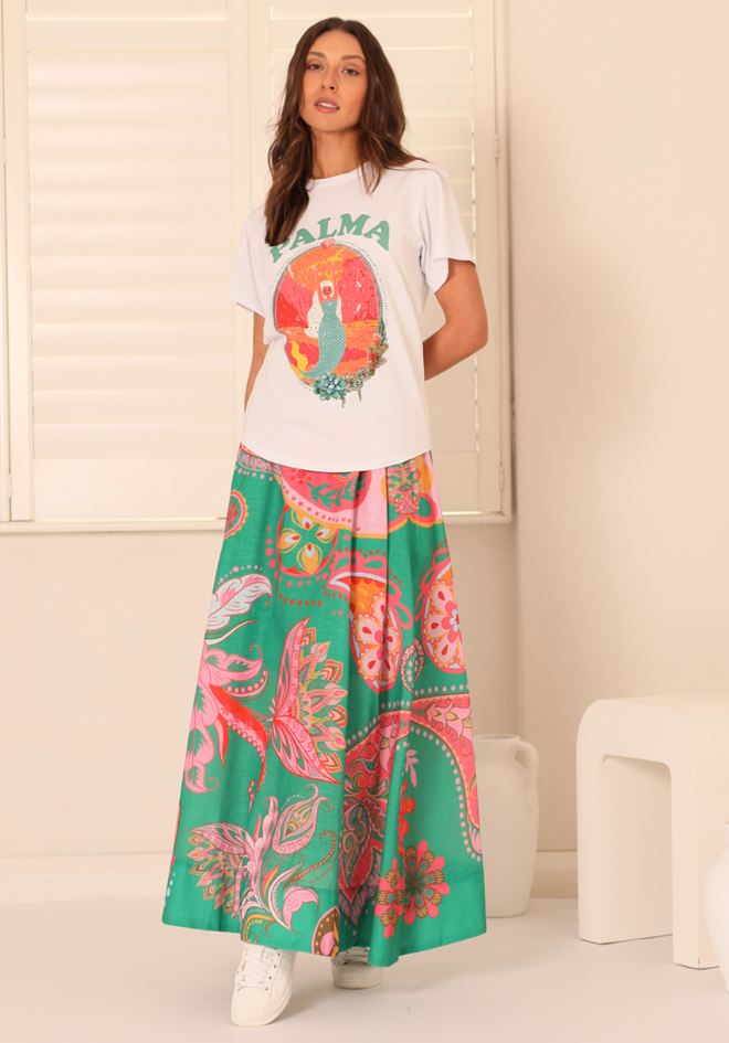 Woman wearing a white t-shirt with a colorful design and a long green skirt with pink patterns in a minimal indoor setting.