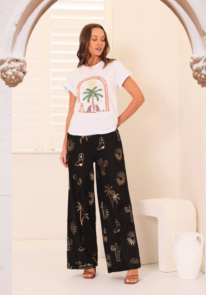Woman wearing a white t-shirt with a palm tree design and black patterned pants in a bright room.