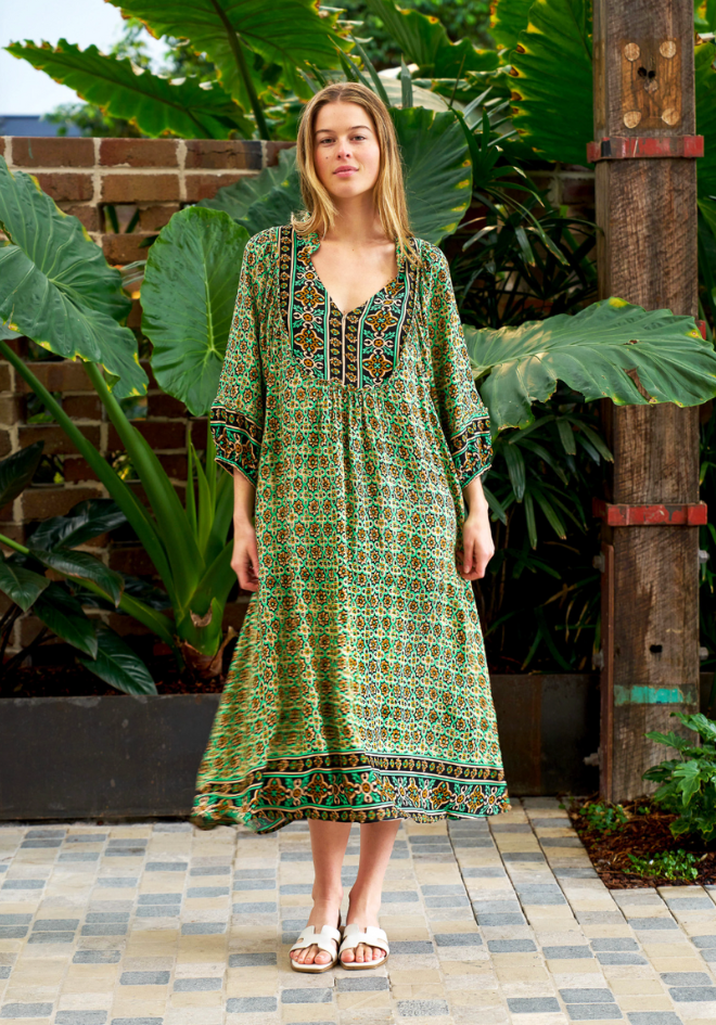 Woman wearing a green patterned dress standing outdoors with plants and a brick wall in the background.