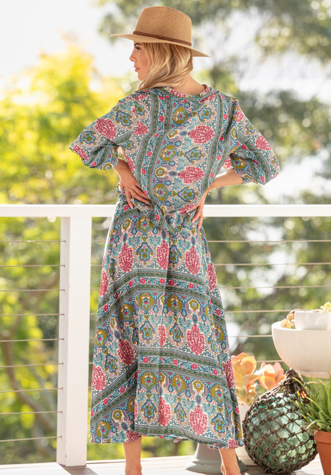 Woman in a patterned dress and hat standing on a balcony with greenery in the background