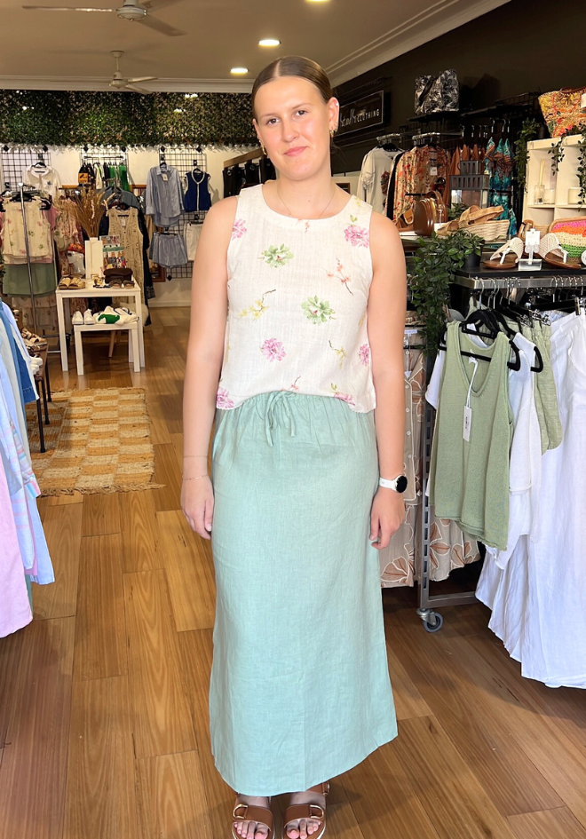 Woman standing in a clothing store wearing a floral top and light green skirt.