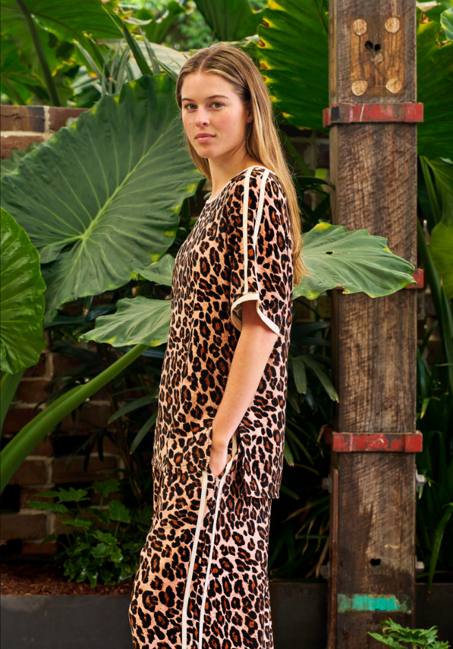 Woman wearing a leopard print dress standing in front of large green leaves and a wooden post.
