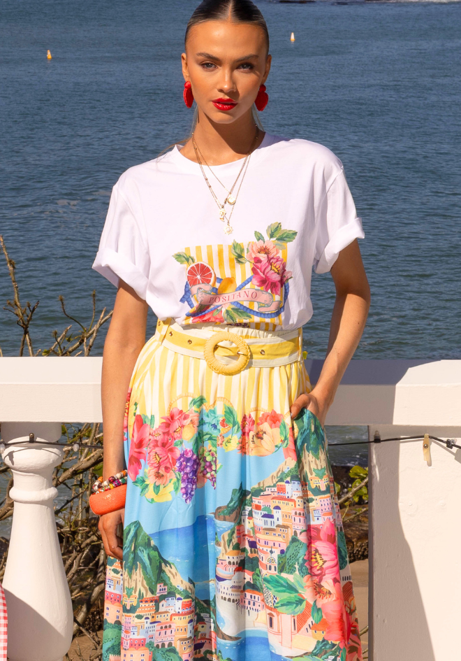 Woman wearing a white t-shirt with a colorful graphic and a patterned skirt by the water.