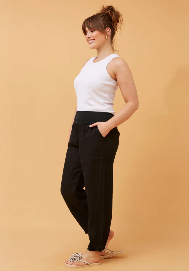 A model wearing black linen pants with a wide elastic waistband and a white top, standing against a beige background.