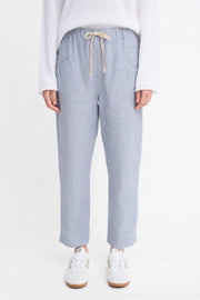 Luxe Pants by Little Lies — close-up front view of chambray blue linen pants with elasticated waist and cream drawstring tie, styled with a white long-sleeve top and white sneakers, available in Australia at Kat and Ko Clothing