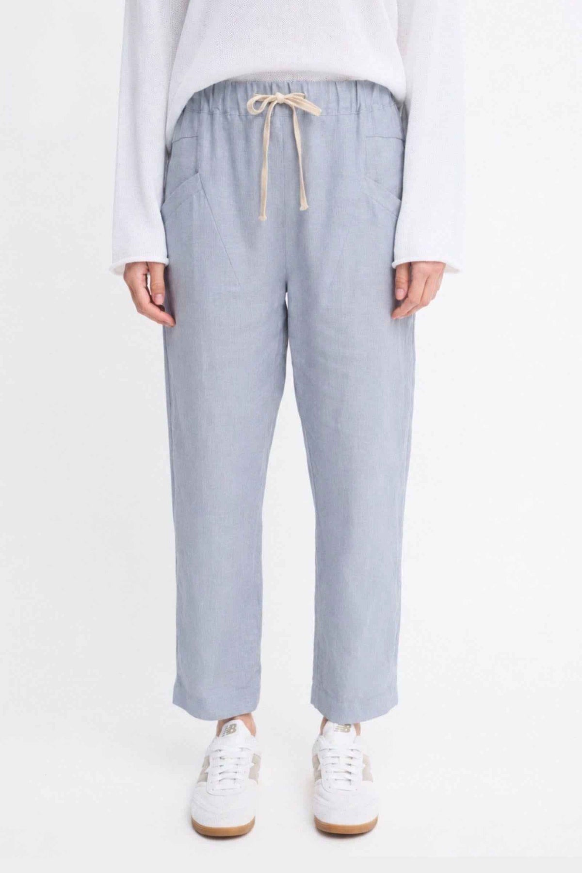 Luxe Pants by Little Lies — close-up front view of chambray blue linen pants with elasticated waist and cream drawstring tie, styled with a white long-sleeve top and white sneakers, available in Australia at Kat and Ko Clothing