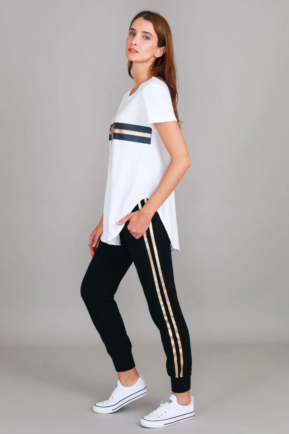 3rd Story The Label Jill Gold Star White T-Shirt — side profile showing the longline hi-lo hem and navy and gold stripe graphic of the white tee, styled with black and gold side-stripe joggers and white Converse sneakers, available in Australia at Kat and Ko Clothing
