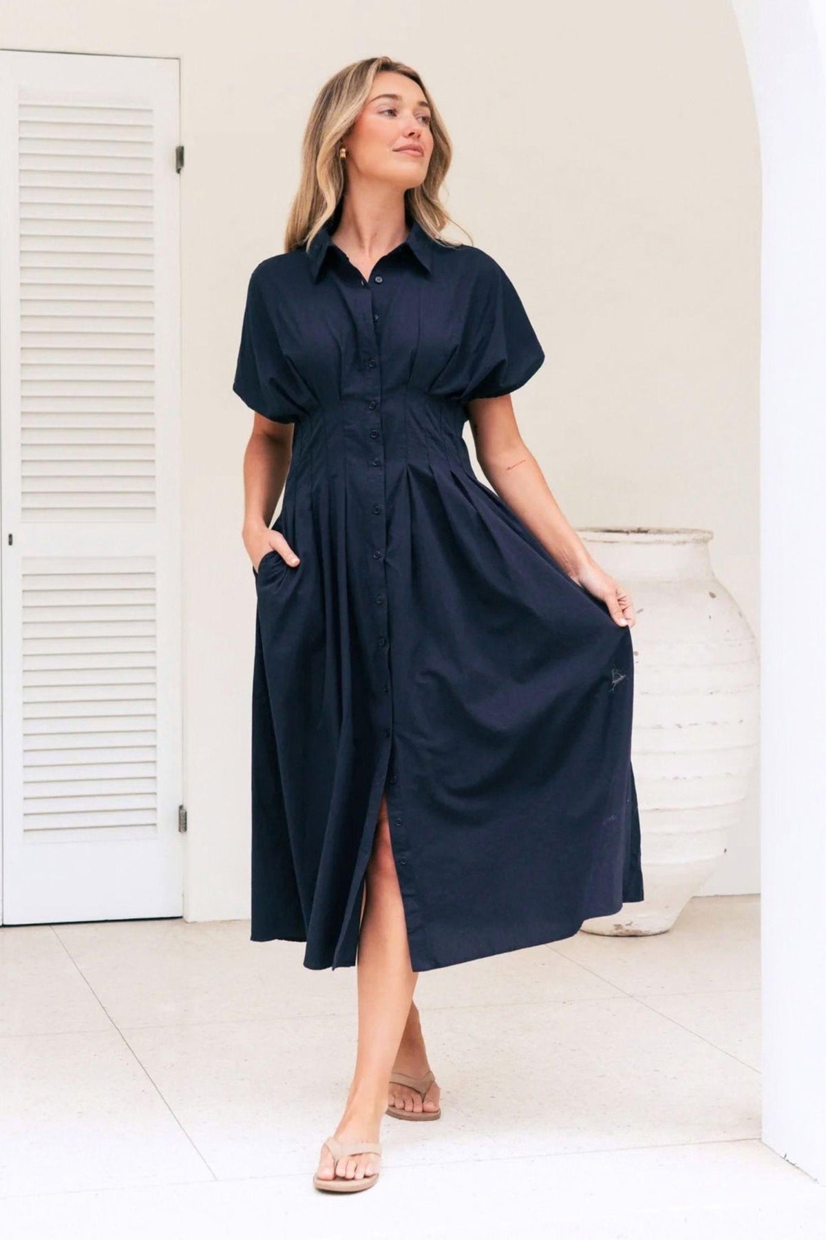 Azure Weekend Midi Dress by Label of Love, front walking view showing the navy button-front dress and flowing pleated skirt, styled with tan flat sandals. Available in Australia at Kat and Ko Clothing.
