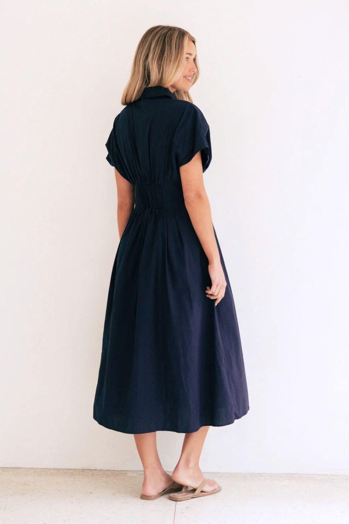Azure Weekend Midi Dress by Label of Love, angled back view showing the navy dress's collar, short sleeves and pleated midi skirt, styled with tan flat sandals. Available in Australia at Kat and Ko Clothing.
