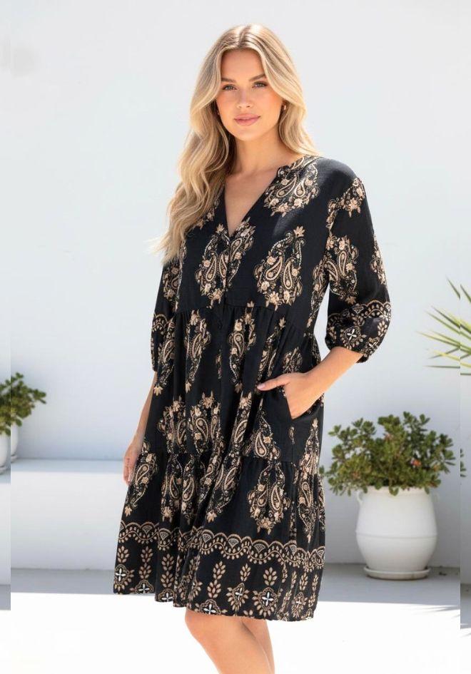 Miracle Carmody Dress — full-length front view of black and gold paisley print tiered dress with V-neckline, 3/4 sleeves and scalloped border hem, styled outdoors with hand in pocket, shop in Australia at Kat and Ko Clothing