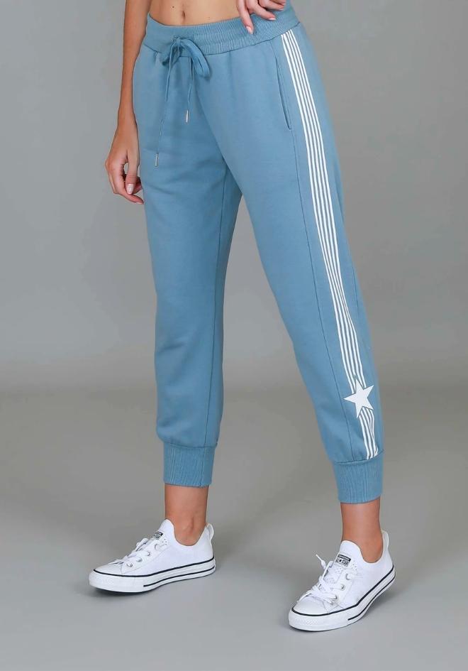 3rd Story The Label Britney Star Stripe Pants — angled front view of steel blue cotton jogger pants with white side stripe and star motif, smocked elastic waistband with drawstring, styled with white Converse sneakers, shop in Australia at Kat and Ko Clothing