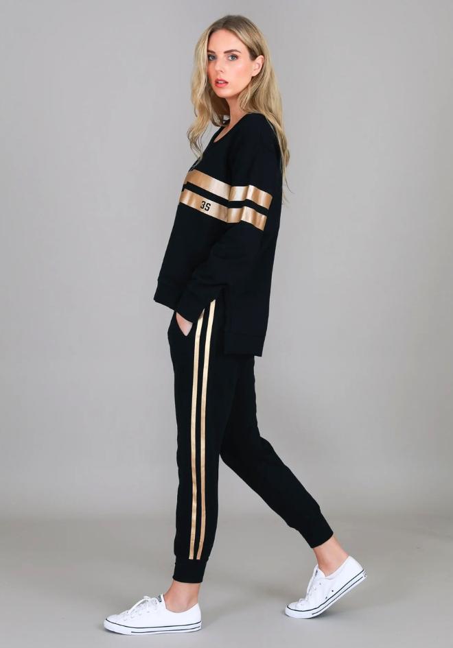 3rd Story The Label Gabby Stripe Jogger Black — full-length side view of black tapered jogger with gold metallic side stripes, styled as a matching set with black and gold stripe sweater and white sneakers, shop in Australia at Kat and Ko Clothing