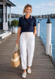 Sailor Knitted Top in Navy by Little Lies — front view of navy knit polo top with red and white stripe collar detail, styled with white linen pants, loafers and woven tote bag at a waterfront setting, available in Australia at Kat and Ko Clothing