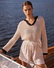 Woman standing on a boat wearing a beige crochet top and white shorts with a blurred water background