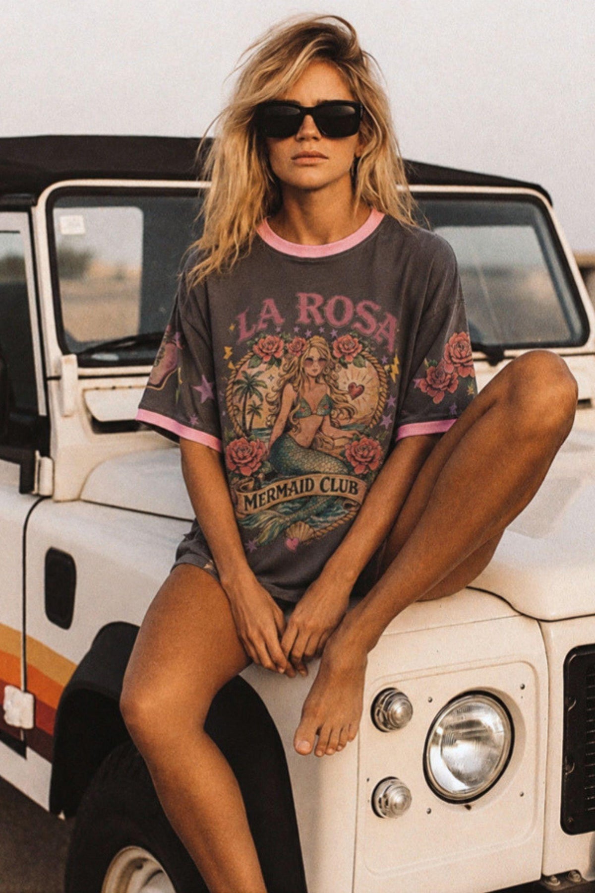 Coco Cartel La Rosa Tee Pink — outdoor lifestyle shot of the charcoal La Rosa Mermaid Club oversized graphic tee with pink ringer trim, worn casually seated on a vintage 4WD, available in Australia at Kat and Ko Clothing