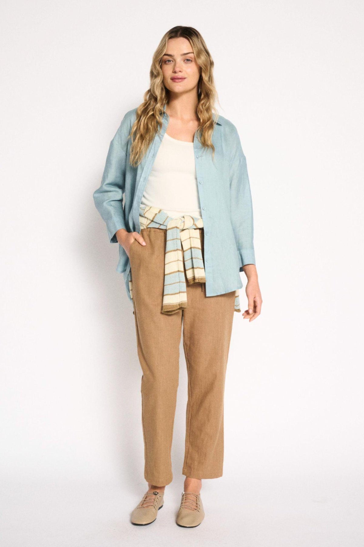 Boyfriend Shirt Aqua Haze Little Lies — full-length lifestyle shot of aqua haze linen shirt worn open with a stripe knit jumper tied at the waist, styled with tan straight-leg trousers and beige loafers, shop in Australia at Kat and Ko Clothing