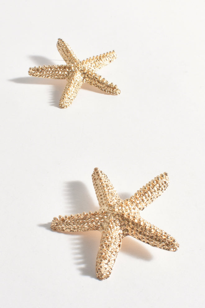 A pair of gold-colored metal starfish earrings with a textured surface, displayed against a white background.