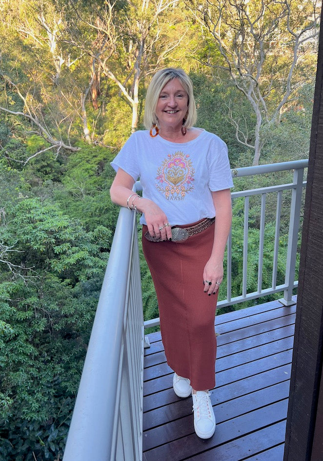 A woman standing on a balcony wearing a rust-colored knit skirt with elastic waist, paired with a white top with a graphic print and beige shoes.
