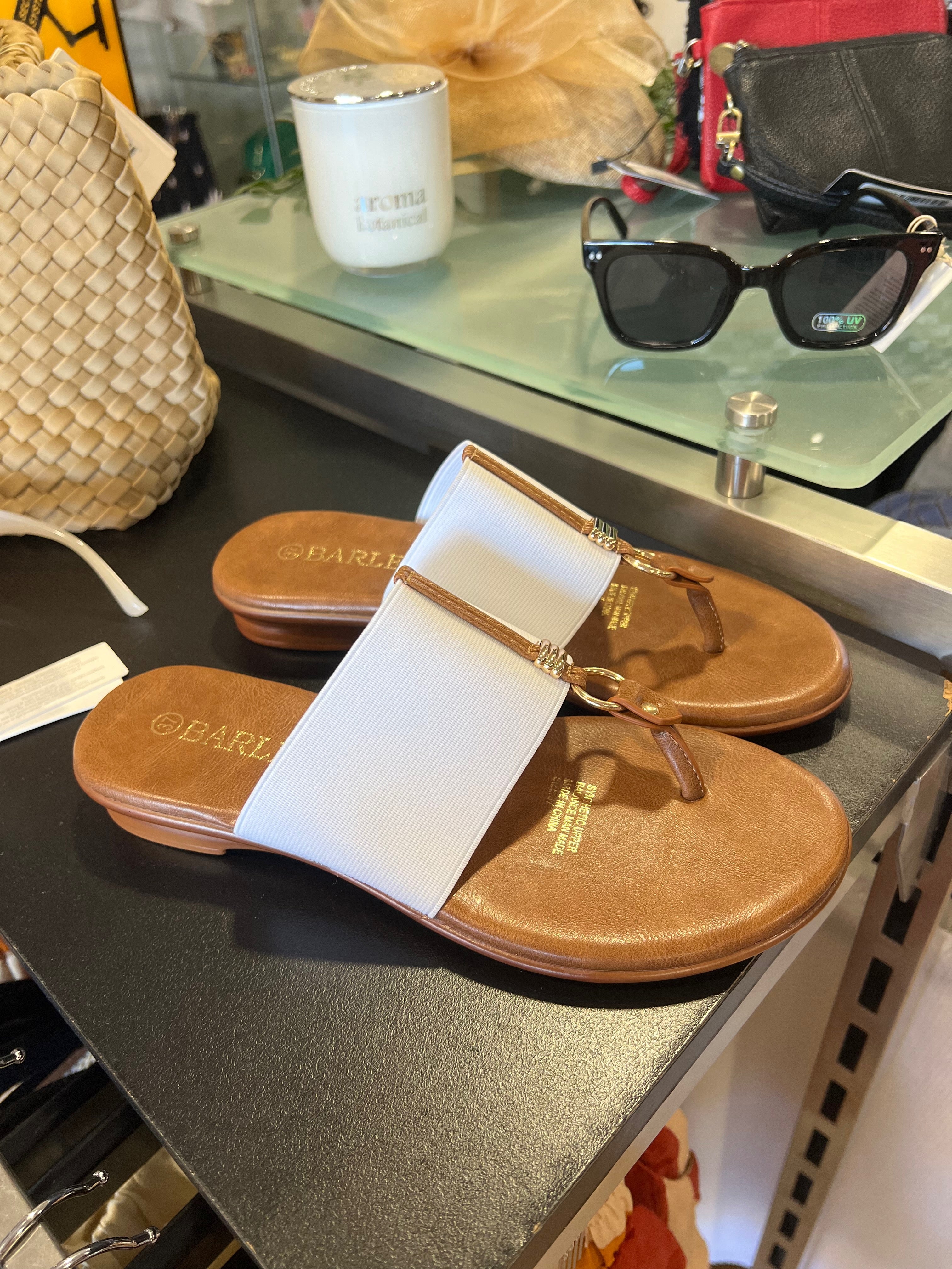A pair of white sandals with brown straps and a label attached, displayed on a glass shelf in a store.