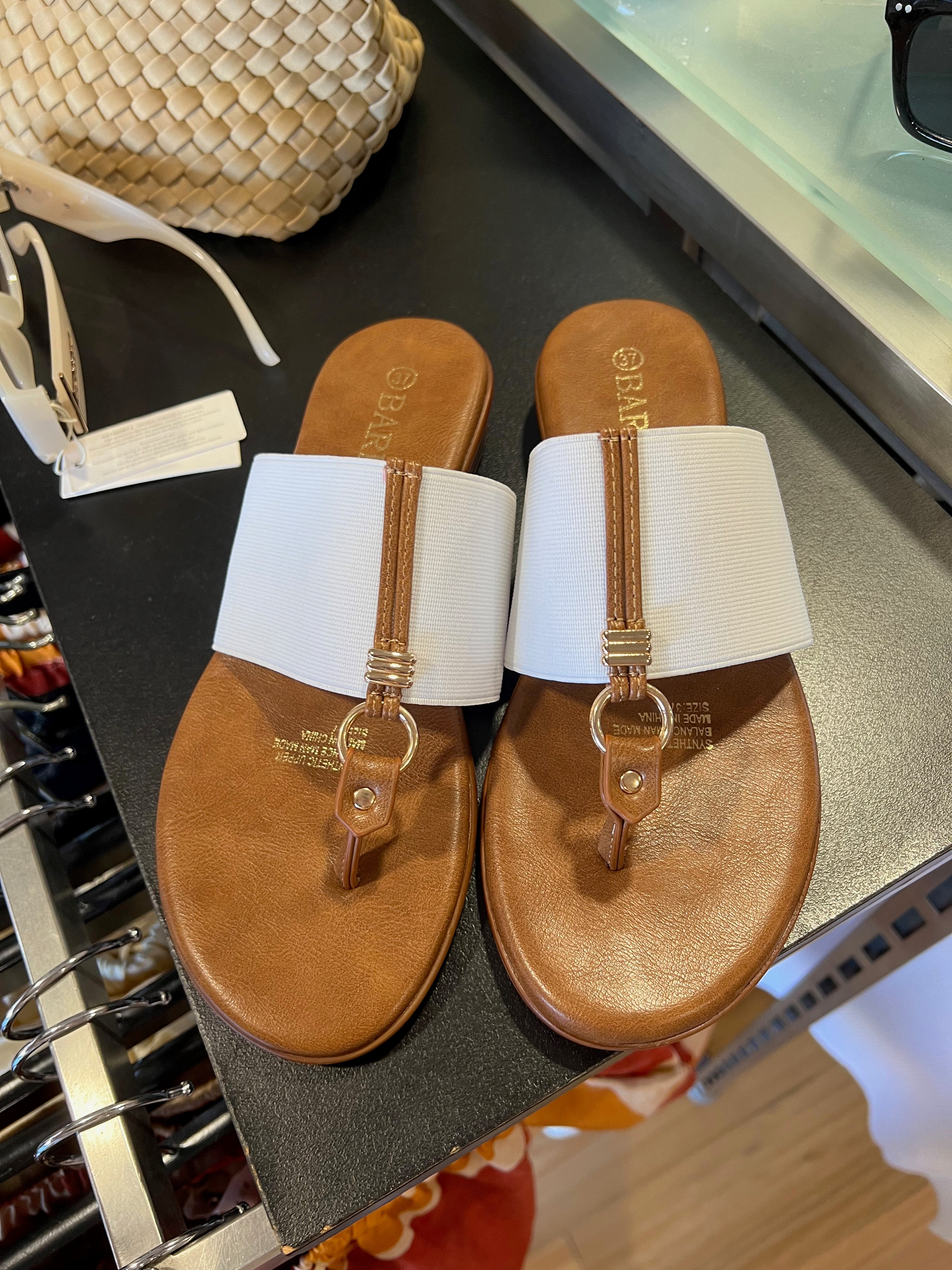 A pair of white sandals with brown straps and a label attached, displayed on a glass shelf in a store.