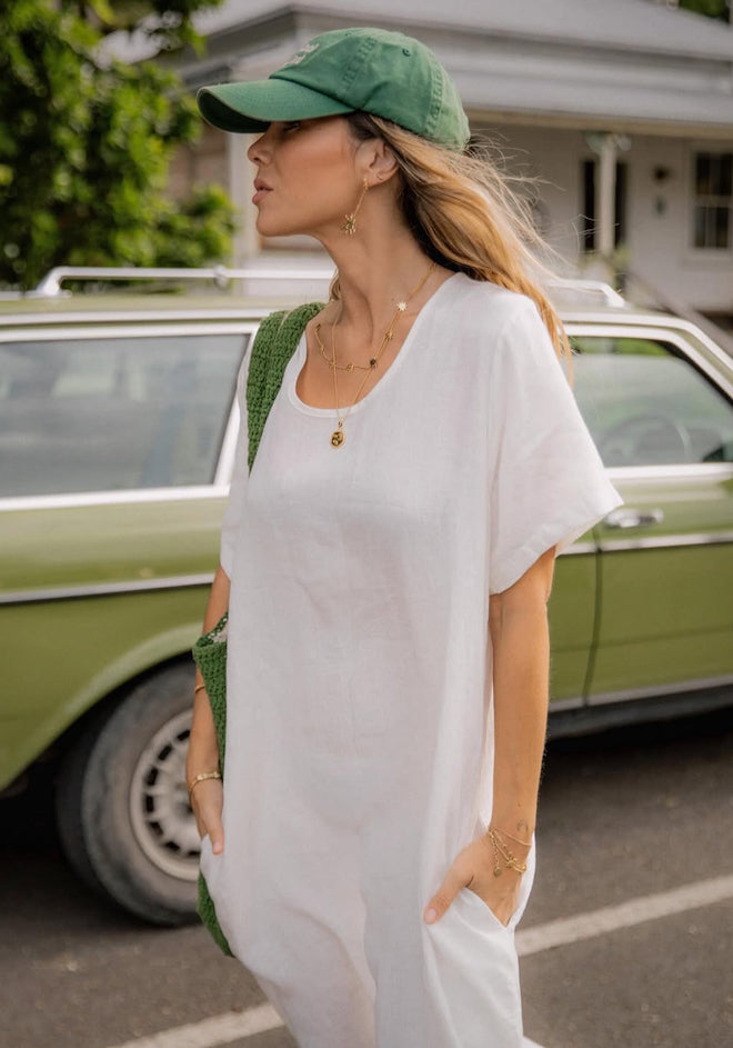 A woman wearing a white t-shaped dress with round neckline and side splits, standing by a green car. She is also wearing a green hat and has a bag with her.