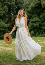 A woman wearing a white, floral print dress with a wide-brimmed hat, standing in a field with trees in the background.