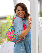 A person wearing a denim dress is showcasing a vibrant multicolored crossbody bag with a geometric pattern and a pink strap.