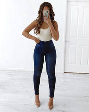 A person wearing dark blue pull-on jeans with knee detail, paired with a white top, standing in a room with light-colored walls.