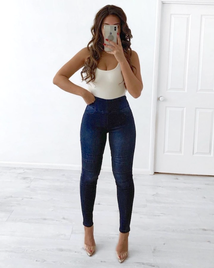 A person wearing dark blue pull-on jeans with knee detail, paired with a white top, standing in a room with light-colored walls.